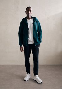 Teal zip-up hoodie over a white t-shirt, paired with black joggers and white sneakers. The hoodie features a simple design and pockets.