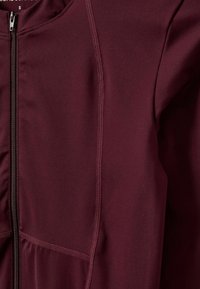 Burgundia värvi sportlik jakk, millel on lukuga eesmine osa, sile tekstuur ja nähtavad õmblused, ning vöökoht on kaunistatud volditud kangaste detailidega.