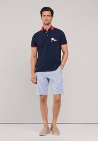 Granatowa koszulka polo z czerwono-białymi akcentami oraz kieszonką w nadruk żaglówki, w zestawie z jasnymi spodniami w prążki i beżowymi loafersami.