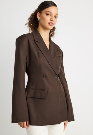 Młoda kobieta z kręconymi włosami nosi ciemnobrązową, oversize'ową, dwurzędową marynarkę i białe spodnie na jednolitym tle.