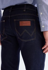 Vyras dėvi tamsiai mėlynas Wrangler džinsus ir šviesiai mėlyną marškinius su sagomis, rodo užpakalinę kišenę su odine prekės ženklo etikete.