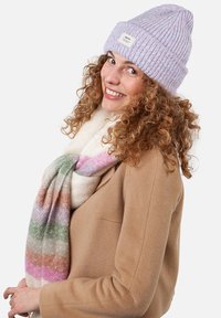 Lavendelstrikbeanie, beige tørklæde med flerfarvede striber, lyst brunt frakke, krøllet hår, smilende udtryk.