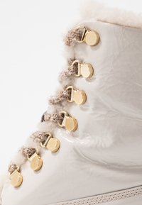 Botín de tobillo beige brillante con un cuello de tela texturizada, herrajes dorados y un distintivo detalle de cordones con lazos y ganchos alternos.