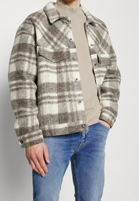 Brun och krämrandig kavaj av tjock ull, med krage, knappstängning och två bröstfickor. Bärs över en beige skjorta.