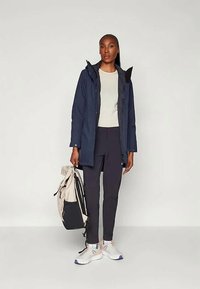 Tumedast veekindel jakk, millel on eemaldatav kapuuts, millele on lisatud heledast kreemjas toonis särk ja tume püksid. Käes on beez ja must seljakott ning jalas on sneakersid.