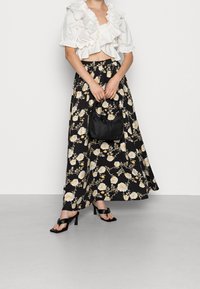 Kvinna som bär en vit volangcropp och en lång svart blommig kjol, håller en svart handväska och står i svarta klacksandaler.