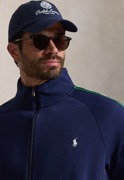 Άνδρας που φοράει σκούφο Ralph Lauren σε ναυτικό μπλε χρώμα, σκούρα γυαλιά ηλίου και μπλε μπουφάν με φερμουάρ με λευκό λογότυπο παίκτη πόλο στο στήθος.