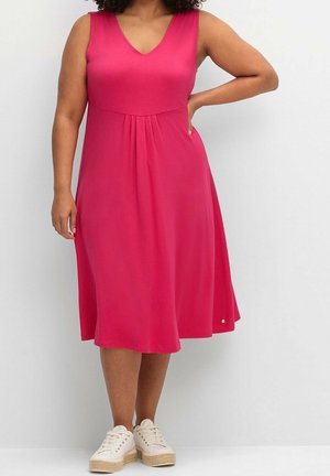 Femme portant une robe rose sans manches longueur genou avec des plis, debout avec une main sur la hanche et des baskets compensées blanches.