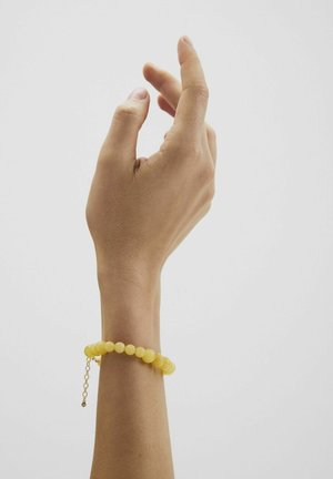 Mano derecha con dedos ligeramente doblados que lleva una pulsera amarilla de cuentas sobre un fondo blanco liso.