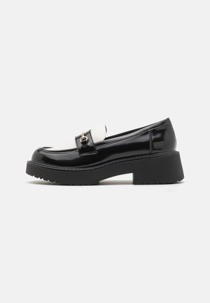 Black patent leather loafers with a white vamp, featuring a gold buckle detail and a thick black textured platform sole.