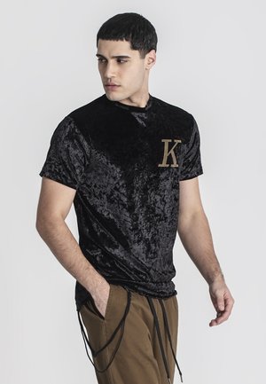 Černé sametové tričko s kulatým výstřihem a krátkými rukávy, s detailem zlatého písmene "K" na levé části hrudi.