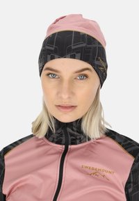 Zwarte en roze beanie met een geometrisch patroon, gemaakt van een lichte, rekbare stof. Heeft een gouden logo langs de rand.