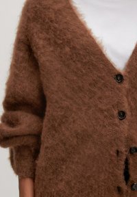 Bruine gebreide cardigan van pluizig materiaal, met een diepe V-hals, lange mouwen en vier zwarte knopen aan de voorkant.
