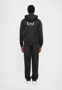 Svart träningsdräkt med huva, med "EA7 Emporio Armani"-logotyp på baksidan. Slät tyg, avslappnad passform, elastiska ärmslut och midja. Vita sneakers.