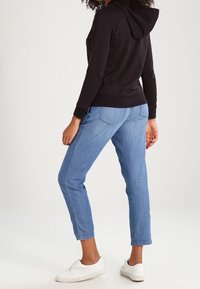 Sweat à capuche noir avec capuche, porté avec un jean bleu clair légèrement fuselé et des baskets blanches sans lacets, vu de dos.