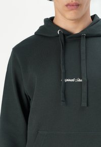 Donkergroene hoodie van zacht materiaal met een voorste kangoeroezak, een capuchon met trekkoord en geborduurde tekst op de borst.