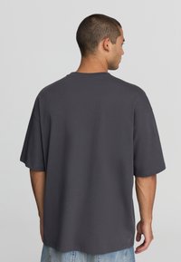 Mężczyzna z krótkimi włosami, ubrany w luźny ciemnoszary T-shirt i jasnoniebieskie dżinsy, stojący tyłem na jednolitym jasnoszarym tle.