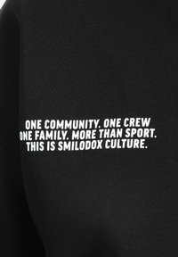 Czarny materiał o prążkowanej teksturze z białym napisem: "JEDNA SPOŁECZNOŚĆ. JEDNA EKIPA. JEDNA RODZINA. WIĘCEJ NIŻ SPORT. TO JEST KULTURA SMILODOX."