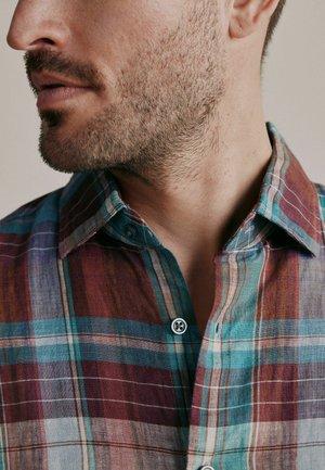 Close-up of a man’s lower face with short beard, wearing a multicolored plaid button-up shirt with blue, red, and brown tones.