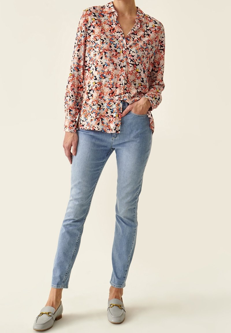 Person som bär en långärmad blommig skjorta med knappar, ljusblå jeans och ljusgrå loafers med guldhästsko-detajl.