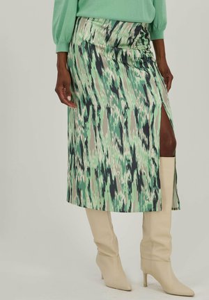 Jupe midi à motif vert avec une fente haute, fabriquée en tissu lisse ; assortie à des bottes beiges jusqu'aux genoux.