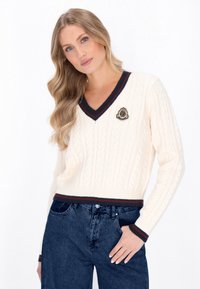 Cremfarbener Kabelstrickpullover mit V-Ausschnitt, der marineblaue und burgunderrote gestreifte Akzente am Kragen und Saum sowie ein dekoratives Patch auf der Brust aufweist.