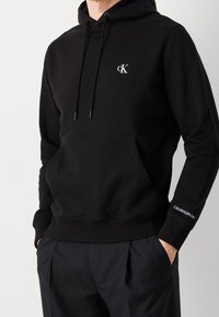 Black cotton hooded sweatshirt featuring a kangaroo pocket, drawstring hood, and logo accent on the sleeve. Smooth texture and regular fit.