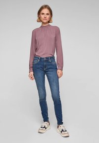 Fialová blůza s nařaseným v�ýstřihem a dlouhými rukávy, v kombinaci s přiléhavými modrými džíny a bílými teniskami s černými a béžovými akcenty.