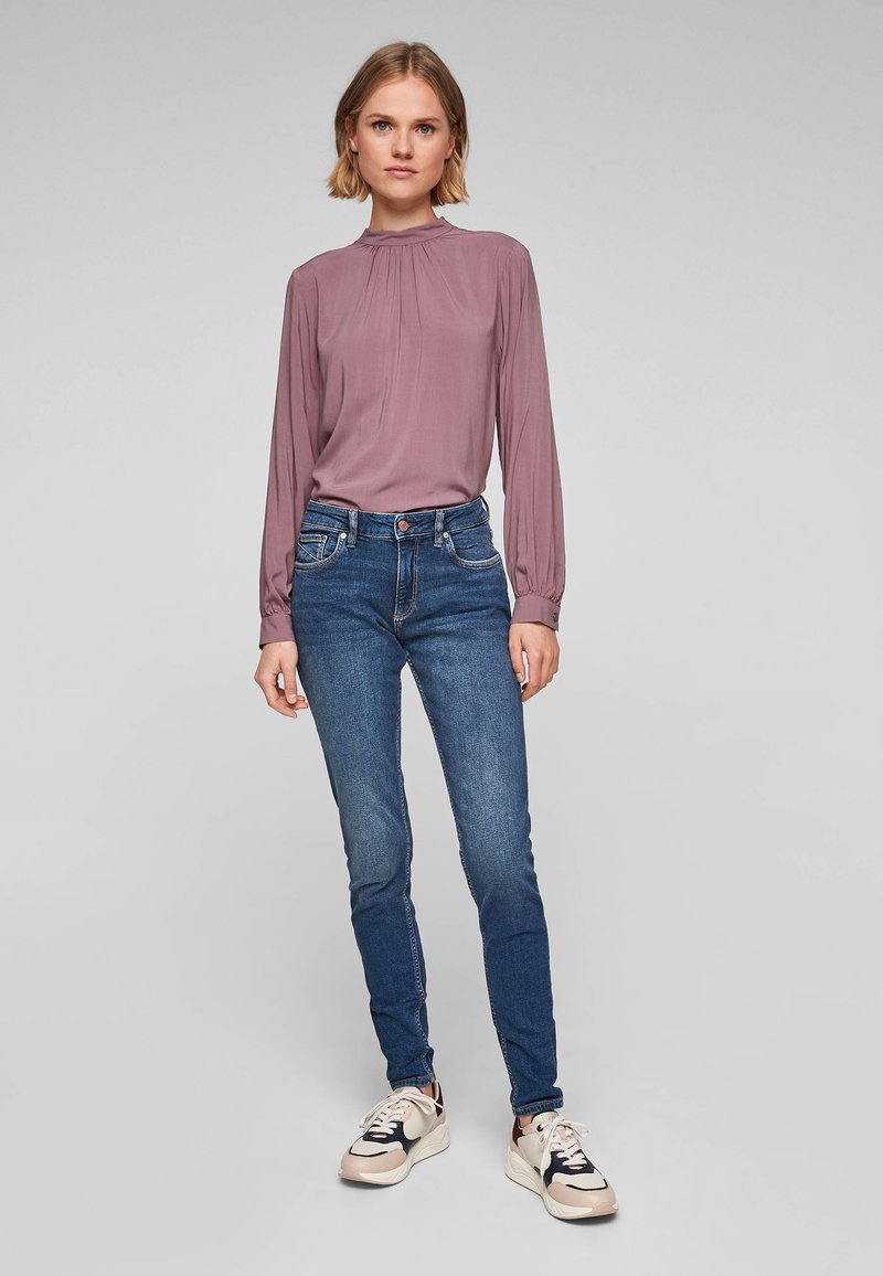 Fialová blůza s nařaseným výstřihem a dlouhými rukávy, v kombinaci s přiléhavými modrými džíny a bílými teniskami s černými a béžovými akcenty.