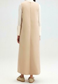 Beige pitkä mekko, jossa on suora leikkaus, korkea kaula-aukko ja pitkät hihat. Sileä kangas ja takakappaleen vetoketjukiinnitys, yhdessä beigejen jalkineiden kanssa.