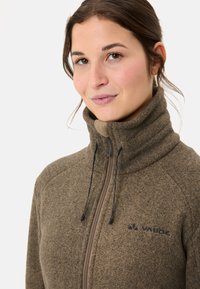 Giacca in pile marrone con colletto alto, zip frontale e cordino nero. Presenta una leggera texture e un logo sul lato sinistro del petto.