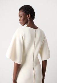 Cream-colored dress with short, wide sleeves, featuring a zip closure at the back and textured vertical stitching along the shoulders.