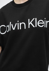 Μαύρο μπλουζάκι με μεγάλο λευκό κείμενο "Calvin Klein" στο στήθος, εμφανίζεται σε άτομο από τον λαιμό έως το μέσο του κορμού.