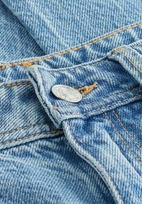 Vaaleansininen farkkukangas, jossa on näkyvää rakennetta; siinä on metallinen nappi ja vahvistettu sauma hieman tummemmassa sinisessä sävyssä.