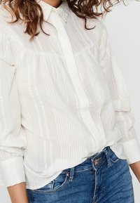 Vit skjorta med vertikala strukturstripor, långa ärmar och krage, i kombination med blå jeans.