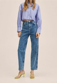 Pantalones vaqueros de tiro alto en azul claro con corte recto, combinados con una camisa de botones corta a rayas en morado y blanco. Sandalias de tacón alto amarillas.