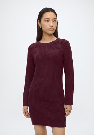 Femme portant une robe en maille bordeaux à manches longues, longueur genou, debout devant un fond blanc, regardant vers sa droite.