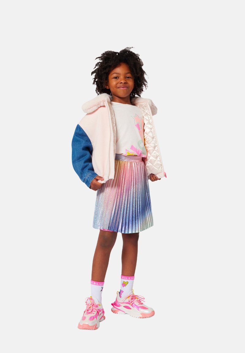 Un bambino che indossa una giacca in tessuto misto rosa e blu, una camicia bianca con stelle colorate, una gonna plissettata iridescente e scarpe da ginnastica rosa.