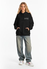 Felpa nera con cappuccio e cordini, tasche frontali e testo bianco. Abbinata a jeans larghi in lavaggio chiaro e scarpe da ginnastica multicolori.