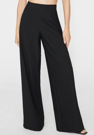 Mujer que lleva pantalones negros de talle alto y pernera ancha, combinados con sandalias negras abiertas, de pie frente a un fondo blanco liso.