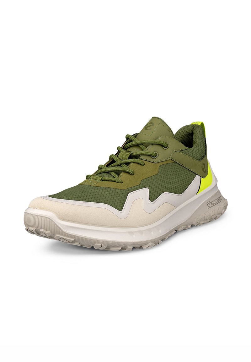 Zapatilla deportiva verde y beige con una parte superior texturizada, paneles de malla y distintivos acentos blancos en forma de ondas. Presenta cordones verdes y un tirador en el talón de color amarillo.