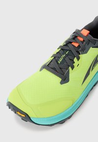 Bottines de course vertes et bleues vif avec des lacets gris foncé, des boucles de lacets orange et une semelle Vibram sur fond blanc.