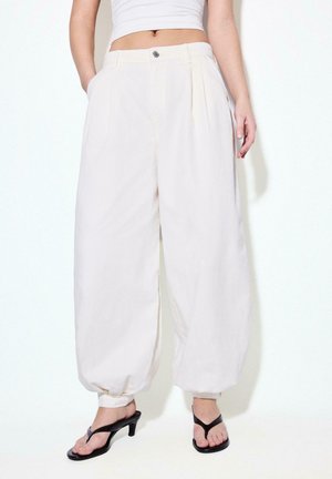 Kobieta nosząca luźne, białe spodnie z wysokim stanem i mankietami przy kostkach, w połączeniu z czarnymi, na obcasie klapkami typu thong oraz krótkim białym topem.