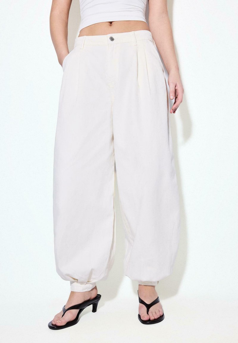 Femme portant un pantalon blanc ample à taille haute avec des chevilles retournées, associé à des sandales à talons noires et un top blanc court.