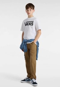 Hvid t-shirt med "VANS"-logo, lyseblå sweater bundet om taljen, brune bukser og blå-sorte sneakers, stående på en lys baggrund.
