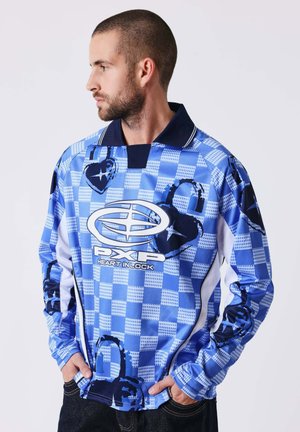 Man met kort haar en baard, gekleed in een blauw geruit overhemd met lange mouwen met zwarte hartvormige slotontwerpen en wit logo, handen in de zakken.