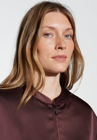 Top in satin bordeaux con scollo rotondo e chiusura con zip frontale. La texture morbida e il design elegante ne esaltano l'aspetto.