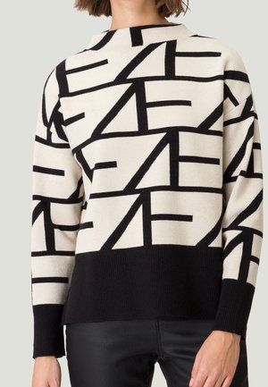 Femme portant un pull crème avec des lignes géométriques abstraites noires audacieuses et un ourlet et des poignets noirs unis, assortis à un pantalon noir.