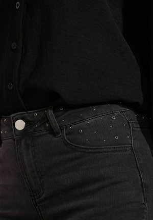 Primo piano di jeans di denim neri con piccoli chiodini metallici rotondi vicino alla vita, abbinati a una camicia nera con bottoni.