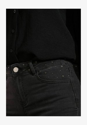 Gros plan sur un jean en denim noir avec de petites clous en métal ronds près de la taille, associé à une chemise noire à boutons.